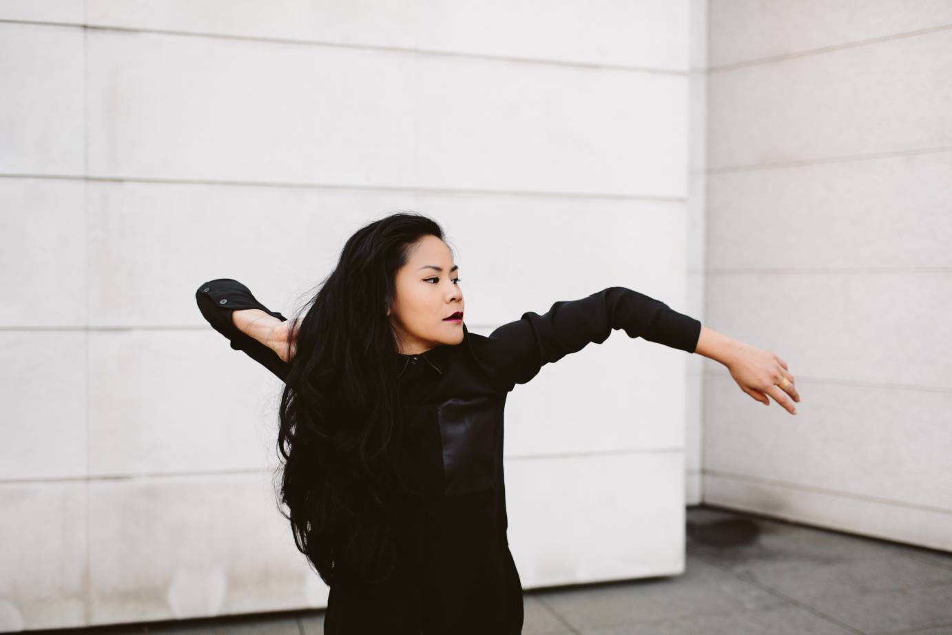 Devin Oshiro, and Asian Woman with long black hair wearing an outfit of all black poses against an all white back ground. She gazes to one side with one arm bent towards her hair and the other arm extended out to where she is looking, but in an angle. her red lipstick pops.  